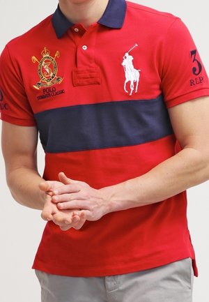 Polo rouge avec une bande horizontale marine, logo brodé et numéro sur la manche. Fabriqué en coton, il présente un col classique et trois boutons.