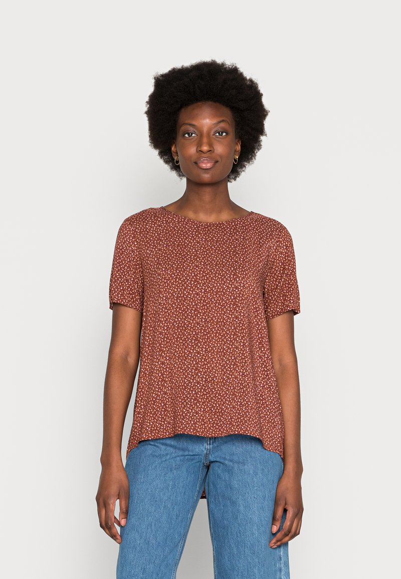Top de manga curta com padrão floral castanho, decote redondo, corte solto e barra ligeiramente curvada. Combinado com jeans de ganga azuis.