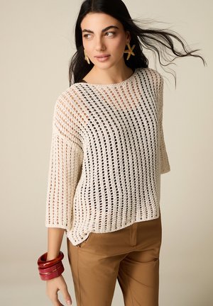 Mujer con cabello largo y negro lleva un suéter blanco de punto abierto, pantalones marrones, pendientes en forma de estrella de mar y pulseras rojas, mirando hacia un lado.