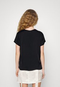 Blusa nera a maniche corte con una texture liscia, caratterizzata da una chiusura a goccia sulla schiena e una vestibilità rilassata, abbinata a una gonna di colore chiaro.