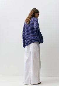 Donna con capelli lunghi castani che indossa una maglia a maniche lunghe a righe blu e marroni e pantaloni bianchi a gamba larga, in piedi davanti a uno sfondo bianco uniforme.