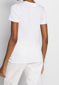 Camiseta blanca de manga corta con un acabado suave y texturizado, que presenta un dobladillo curvado y un sutil detalle de costura en la parte posterior. Combinada con pantalones de color claro.