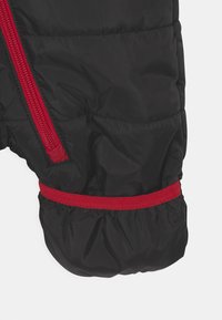 Giubbotto imbottito nero con finitura lucida, caratterizzato da una zip e polsini rossi in contrasto. Accentuato da una banda rossa in vita.