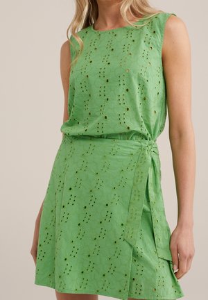 Robe verte sans manches à motif œillet floral et détail lien sur le côté portée par une femme aux cheveux blonds lâchés.