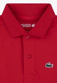 Red polo shirt with a textured fabric, featuring a two-button placket and a green and white Lacoste crocodile logo on the left side.
