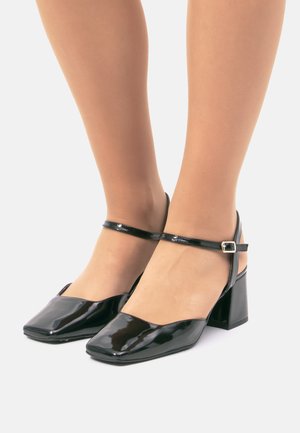 Chaussures à talons blocs en cuir verni noir avec un embout carré et une bride à la cheville réglable. Texture lisse et détails de design minimalistes.
