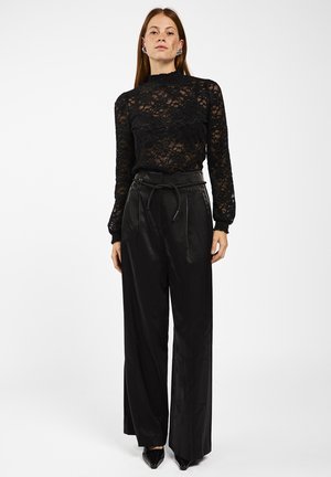 Vrouw in een doorschijnende zwarte kanten top met lange mouwen, hoge taille brede zwarte broek met een riem en zwarte puntige schoenen.
