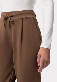 Hnědé kalhoty s vysokým pasem a sklady, vyrobené z jemné látky. Obsahují šňůrku s kovovými akcenty a boční kapsy.