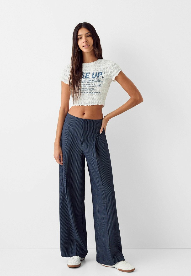 Top bianco cropped con texture arricciata e lettering blu, abbinato a pantaloni a gamba larga a righe blu navy e sneaker bianche.
