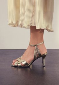 Metallic-goldfarbene Sandaletten mit hohen Absätzen, einem Riemendesign, offenem Zehenbereich, Knöchelriemen und einer glänzenden, glatten Oberfläche. Akzentuiert mit überkreuzten Details.