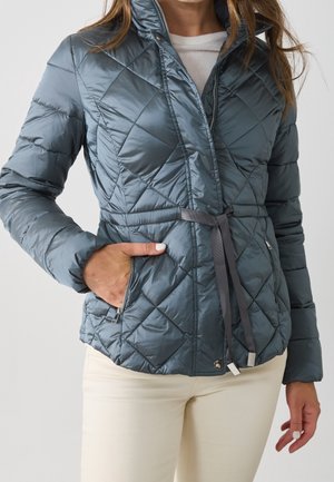 Femme portant une veste matelassée bleue avec une taille cintrée par une ceinture et des poches à glissière, associée à un pantalon beige clair et un haut blanc en dessous.