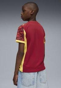 Röd och gul kortärmad tröja med texturerade röda mönstrade ärmar. Har en rund halsringning och en logotyp på axeln. Jeansshorts syns.