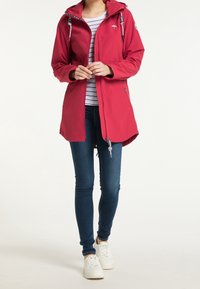 Giacca impermeabile rossa dal design aderente, con cappuccio regolabile con coulisse, fodera interna a righe e bottoni a pressione, abbinata a jeans scuri e sneaker bianche.