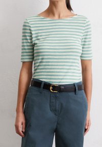 Mujer vistiendo una blusa de rayas verde menta y blanca de mangas cortas metida dentro de pantalones verde azulado oscuros con un cinturón negro con una hebilla dorada.