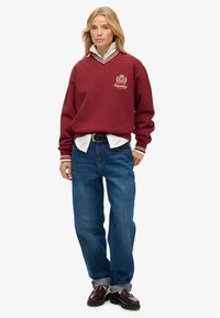 Sudadera burdeos con cuello en V, cuello a rayas blancas y bordado de logo. Combinada con jeans de mezclilla azul y mocasines marrones.