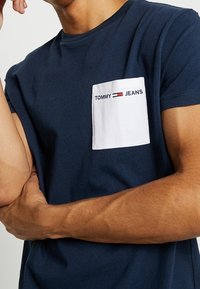 Námořnická modrá bavlněná tričko s bílou náprsní kapsou, na které je logo "TOMMY JEANS" v černém a červeném písmu. Krátké rukávy, design s kulatým výstřihem.
