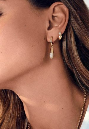 Guldringe med en tåreformet design, der har en struktureret overflade og små krystalaccenter, sat på en models øre.