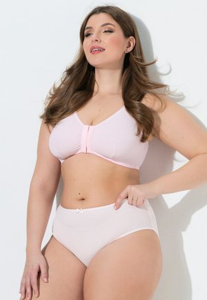 Plus-Size-Frau trägt hellrosa BH mit Frontverschluss und High-Waist-Slips, steht mit der Hand in der Hüfte vor einer weißen Wand.