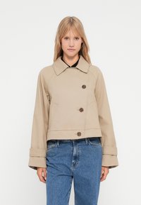 Veste beige courte avec un large col, fermeture à double boutonnage et manches larges. Fabriquée dans un matériau durable au toucher lisse.