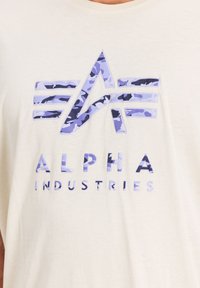 Beige bavlněné tričko s modrým a fialovým maskáčovým logem, pod kterým je v tučném a jasném písmu napsáno "ALPHA INDUSTRIES".