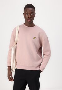 Felpa rosa con collo rotondo e polsini a costine, dotata di un piccolo logo di un uccello ricamato giallo sul lato sinistro del petto. Pantaloni neri e una borsa a tracolla beige.
