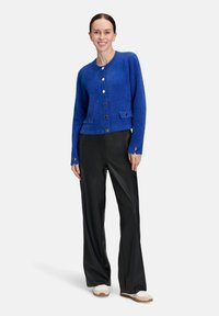 Vrouw in een blauwe, knoopjestrui van fuzzy stof, met zwarte wijde broek en witte sneakers, staand en lachend tegen een neutrale achtergrond.