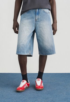 Pantalones cortos de mezclilla con lavado azul claro, hasta la rodilla, con un ajuste relajado y diseño de cinco bolsillos. Combinados con zapatillas rojas y calcetines negros.