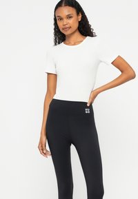 Vit tajt T-shirt med korta ärmar i kombination med svarta högmidjade leggings som har en vit "HUGO"-logotyp på midjebandet.