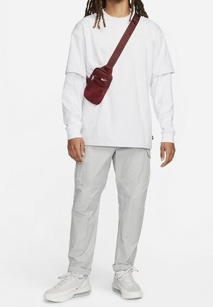 Persoon die een wit shirt met lange mouwen, lichtgrijze broek, witte sneakers en een bordeauxrode crossbodytas met een Nike-logo draagt.