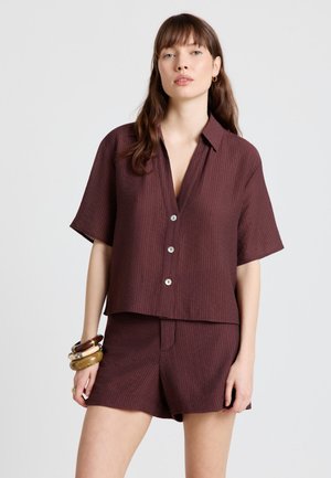 Femme portant une chemise à manches courtes ample de couleur marron foncé avec des boutons et un short assorti, accessoirisée de plusieurs bracelets à son poignet gauche.