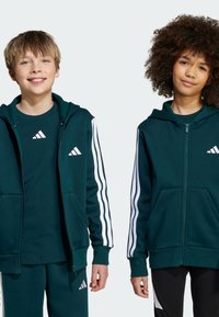 Zelena dukserica na zip s bijelim Adidas prugama na rukavima, prednjim džepovima i kapuljačom. Slim-fit majica u odgovarajućoj boji nosi se ispod.