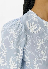 Blusa azul claro con bordado floral blanco, mangas abullonadas y escote en forma de ondas, fabricada con una tela suave de acabado texturizado.