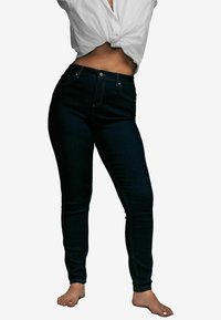Jeans skinny de denim oscuro con cintura alta, costuras doradas en contraste, cinco bolsillos y una textura suave. Llevados con una camisa blanca anudada.