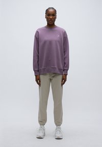 Ανοιχτό μοβ φούτερ με ριμπ μανσέτες, στρογγυλή λαιμόκοψη και διακριτικό λογότυπο. Συνδυασμένο με ανοιχτό μπεζ τζόκερ παντελόνια και λευκά αθλητικά παπούτσια.