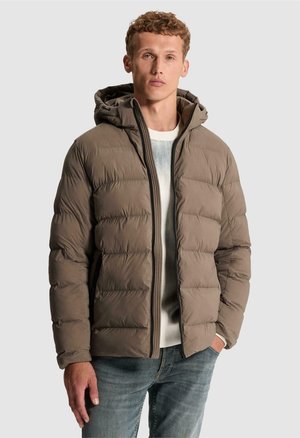 Jeune homme aux cheveux bouclés portant une veste matelassée à capuche marron, un pull blanc et un jean bleu, debout devant un fond uni.