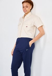 Beez värvi lühikeste varrukatega lühikesed säärised, millel on kaks rinna taskut ja logo. Tumesinised püksid küljetaskutega ja valge Nike swooshi aktsent.
