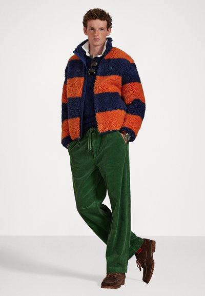 Bársonyos kabát narancssárga és sötétkék csíkokkal, zöld kordbársony nadrággal és barna slip-on cipőkkel párosítva. A modellen napszemüveg van.