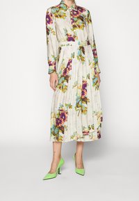 Květinové košilové šaty s dlouhými rukávy, skládáním sukně a živými vícbarevnými květinami na světlém pozadí. Doplněno zelenými podpatky.