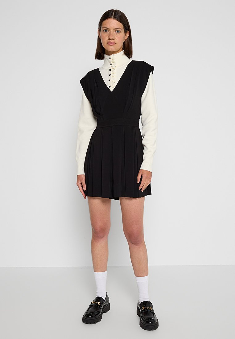 Schwarzes Faltenkleid mit einem weißen hochkragen Top. Mit Knöpfen auf der Vorderseite und schwarzen Loafers ausgestattet. Socken sind über dem Knöchel sichtbar.