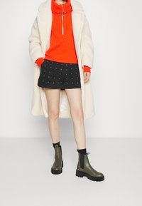 Abrigo de piel sintética beige claro, suéter de cuello alto naranja acanalado, pantalones cortos de mezclilla negra con lunares blancos y botines verde oliva con suelas negras.