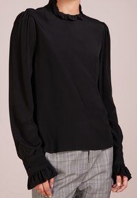 Blusa nera a maniche lunghe con colletto e polsini arricciati, realizzata in tessuto liscio. Indossata con pantaloni a quadri in tonalità di grigio.