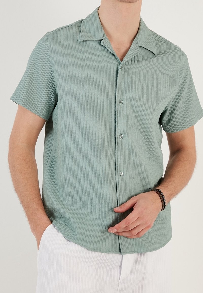 Koszula z krótkim rękawem, zapinana na guziki, w jasnym zielonym kolorze, o fakturowanej, prążkowanej tkaninie. Do wyróżniających cech należy kołnierzyk w kształcie ostrym i subtelny połysk.