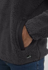 Fleece pullover i mørkegrå med en tekstureret overflade, der har sidelommer og kontrastfarvet sort kant; lille mærke synligt.
