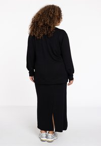 Zwarte oversized sweatshirt met lange mouwen, gecombineerd met een zwarte maxi rok met een split aan de achterkant. Gekleed met zilveren sneakers.