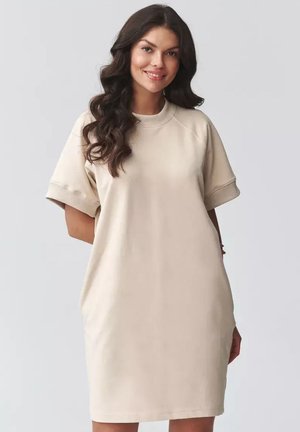 Uśmiechnięta kobieta z długimi falującymi włosami, ubrana w luźną, krótkorękawową beżową sukienkę, stojąca z jedną ręką za plecami.