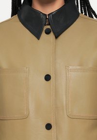 Brūns ādas mētelis ar kontrastējošu melnu apkakli, divām šķēļu kabatām un melnām pogām, gluda tekstūra un redzamas šuves.