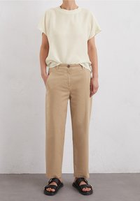 Pantaloni di cotone beige, fit morbido con tasche laterali, abbinati a una maglietta leggera color crema a maniche corte e sandali neri con dettagli dorati.