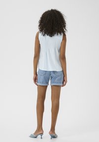 Blouse sans manches bleu clair avec dos froncé, associé à un short en denim roulé, et des chaussures bleu clair à enfiler avec des accents décoratifs.