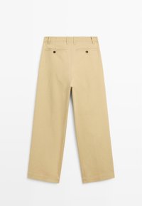 Beigeweit geschnittene Hose mit Gürtelschlaufen und zwei hinteren Taschen, die mit dunklen Knöpfen geschlossen sind, flach vor einem weißen Hintergrund präsentiert.