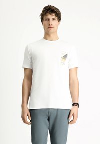 Camiseta de algodón blanca con un estampado en el bolsillo que presenta patrones abstractos en negro y amarillo. Mangas cortas y un diseño clásico de cuello redondo.